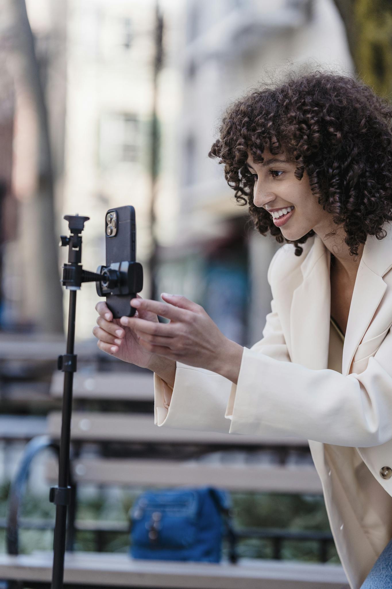 Smiling woman with curly hair uses smartphone and tripod for vlogging outdoors.